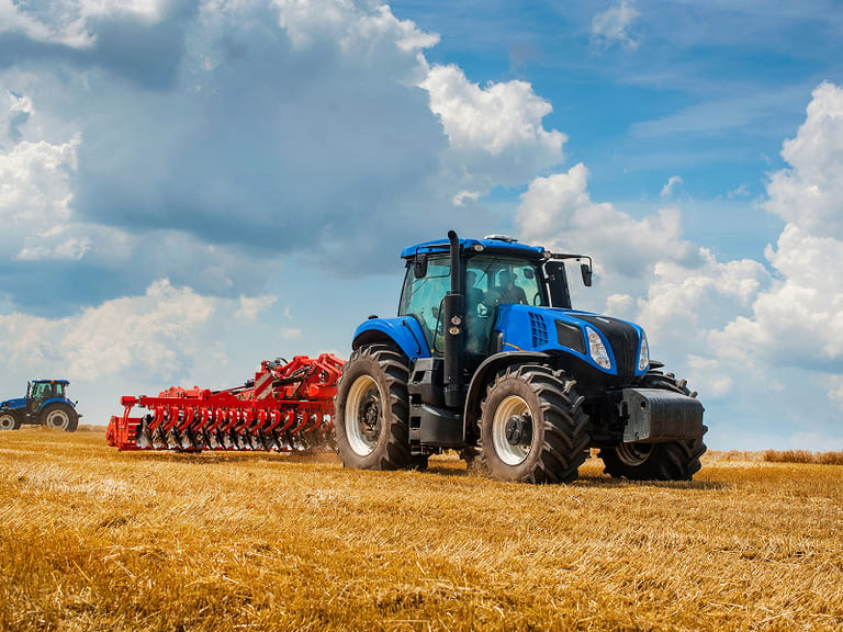 Blue tractor with red cultivator in golden wheat field under blue sky with white clouds.